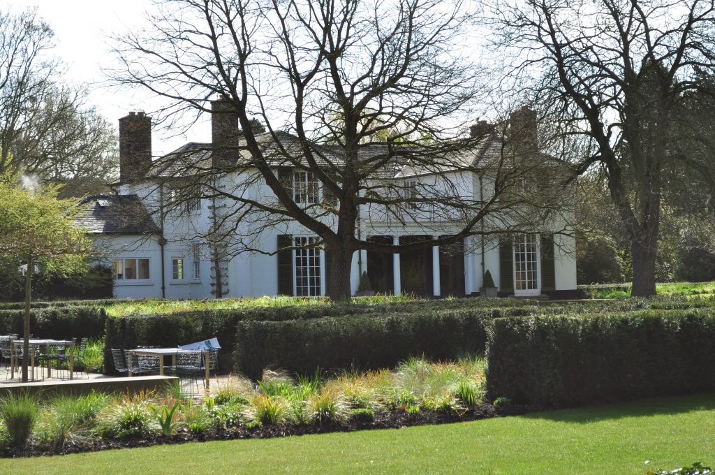 A white house sits next to a cafe, in the middle of a garden with hedges and trees.