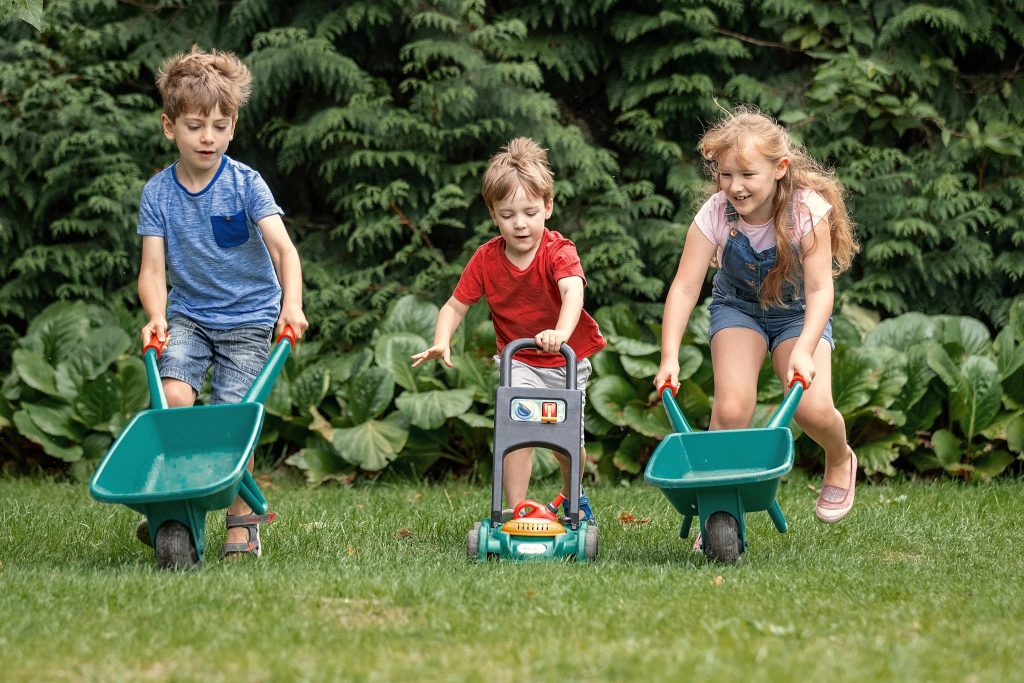 Three children racing across the grass with wheelbarrows.