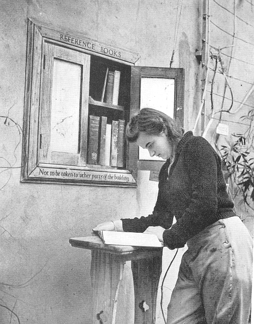 A black and white photo showing a young woman reading a book taken from a small reference library in the wall. She rests the book on a plinth.