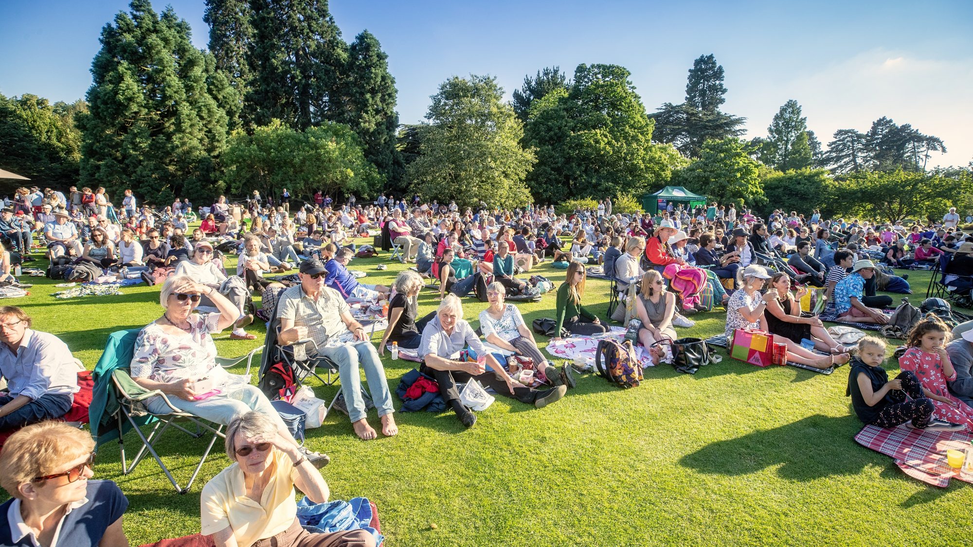 A large crowd sits on a grassy area in a park, enjoying a sunny day. Many people are seated on blankets and folding chairs. In the background, there are tall trees and clear blue skies. The atmosphere appears relaxed and communal.