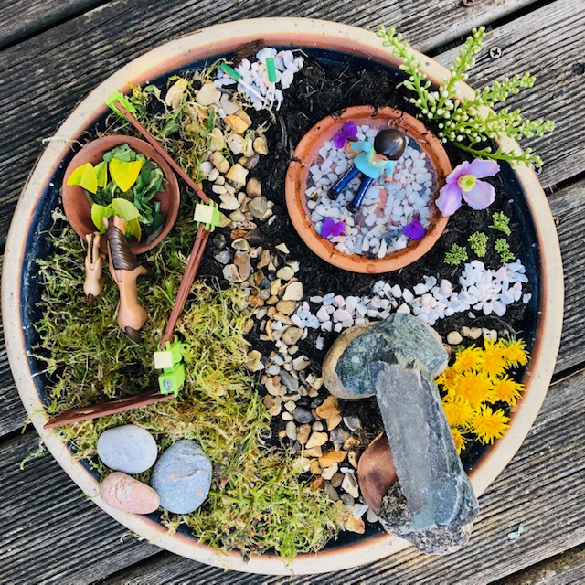Inside a large plant pot is a mixture of stones, flowers, moss and small doll figurines.