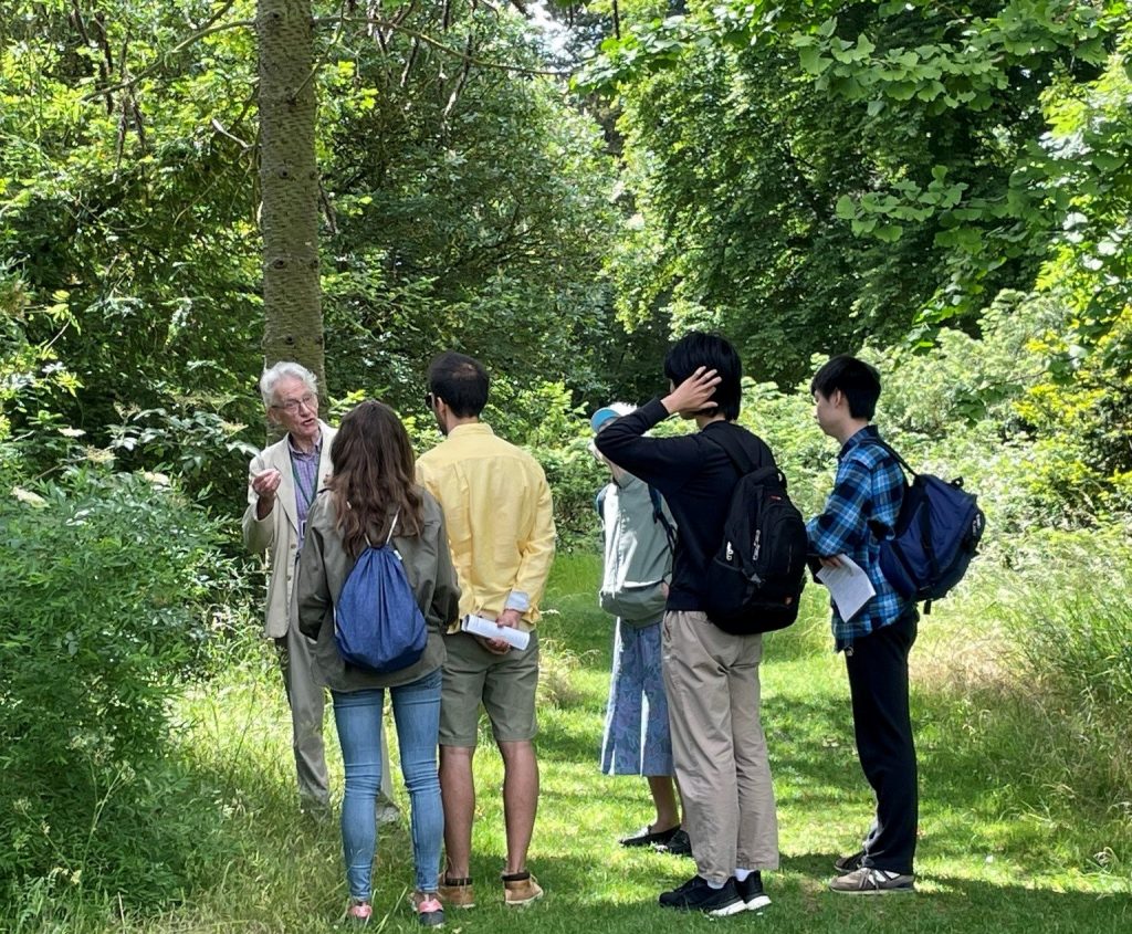 A small group in a woodland clearing listening to a man in a suit.