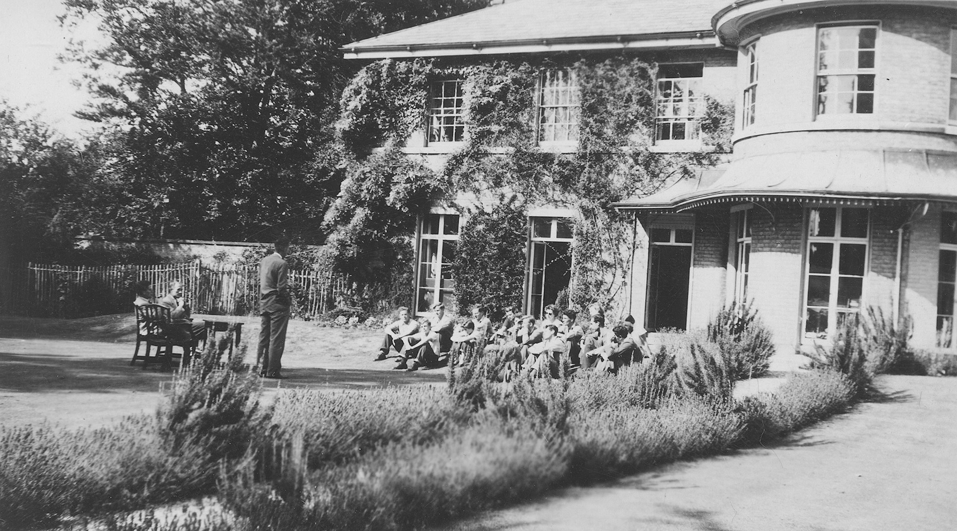 A historical image of a group of people sit together, being taught on the Brookside Lawn.