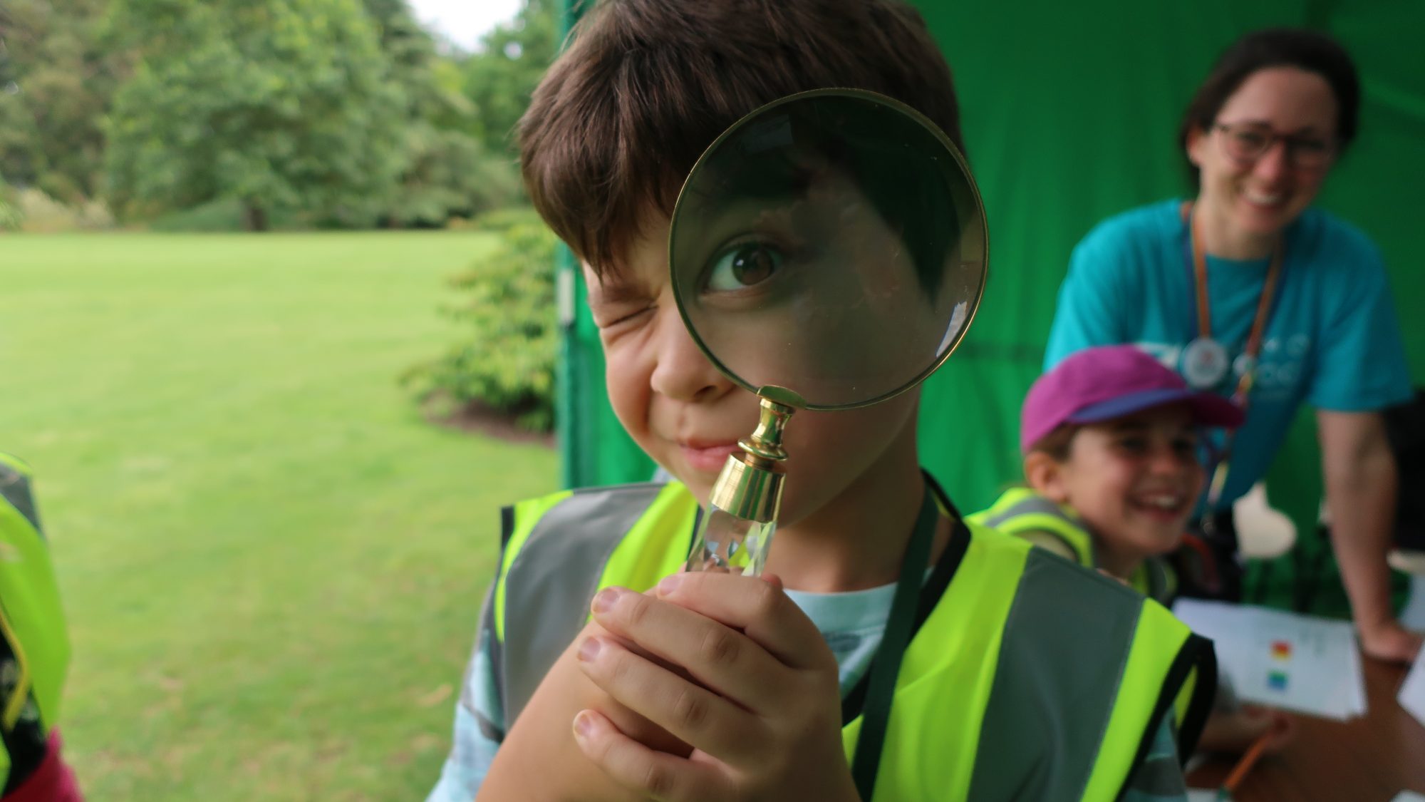 A young boy in a high-vis jacket looks through a magnifying glass at the camera.