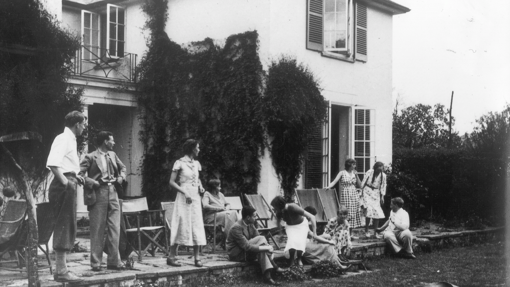 A historic image of Cory Lodge in the Cambridge University Botanic Garden with a group of people gathered in front of it, standing and seated in deckchairs