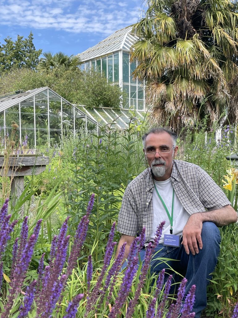 Nabil Ali, Artist-in-residence crouching by some purple flowers with the Glasshouses seen in the background.