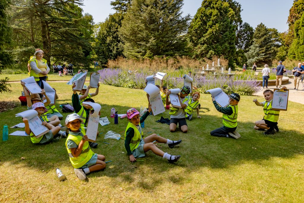 A group of school children in high-vis jackets sit in the shade of a tree with clipboards.