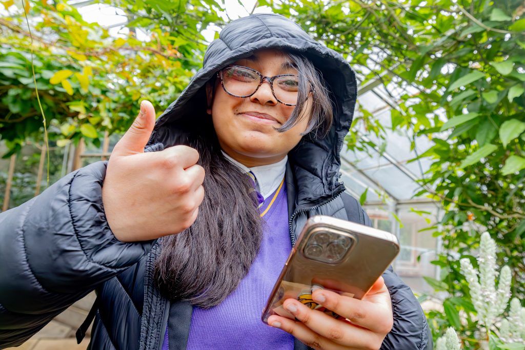 Female high school student in black hooded coat and purple jumper taking photos in the glasshouses