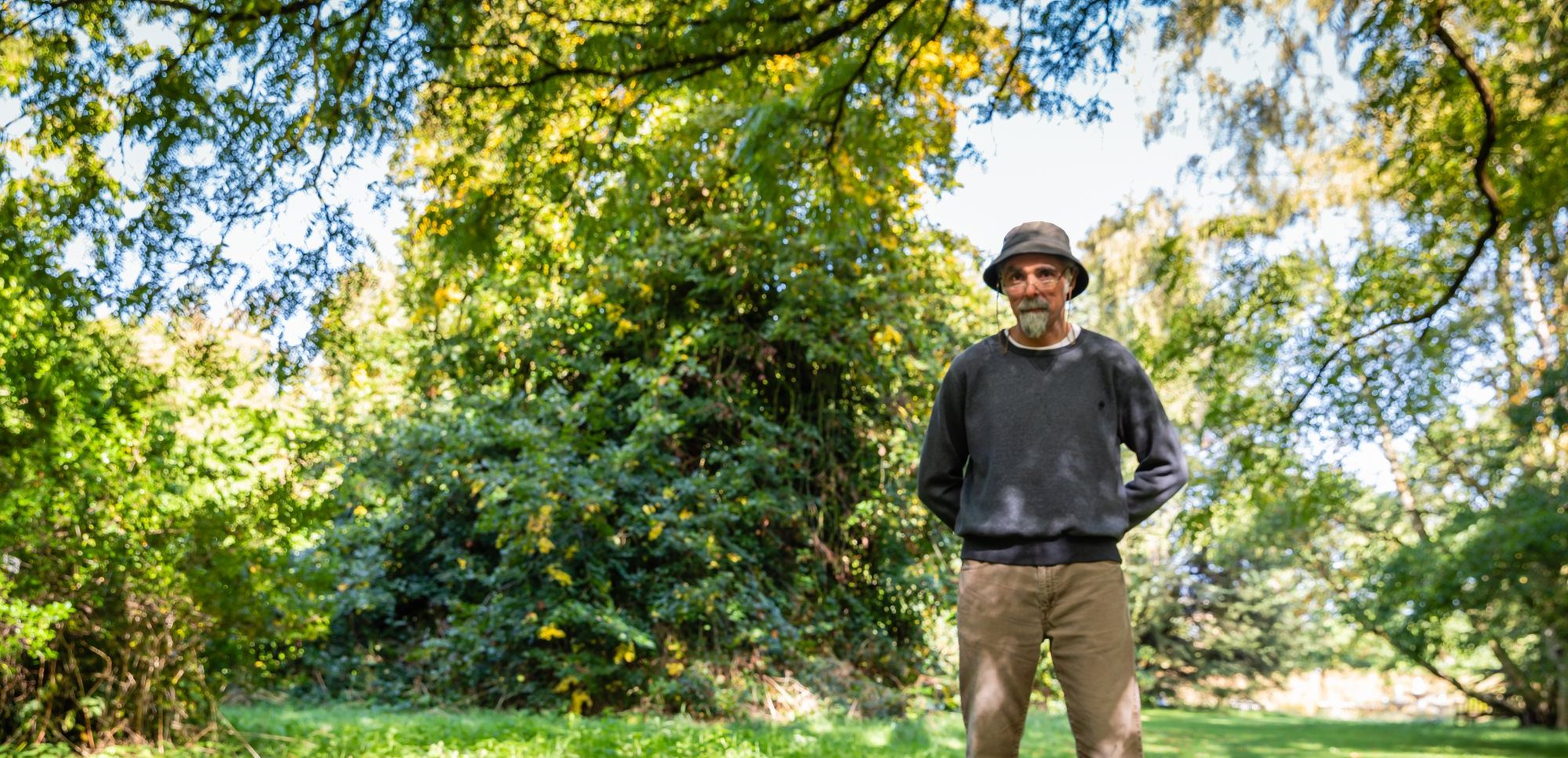 A man in a grey jumper and wearing a bucket hat stands in the middle of a garden under a tree.