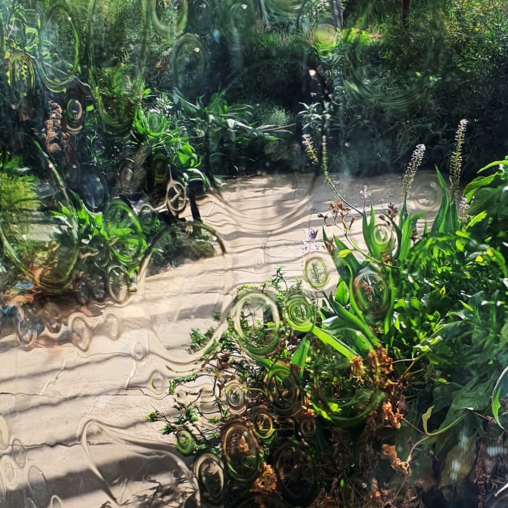 A view into a glasshouse through bubbles in the glass, with greenery visible on the other side.
