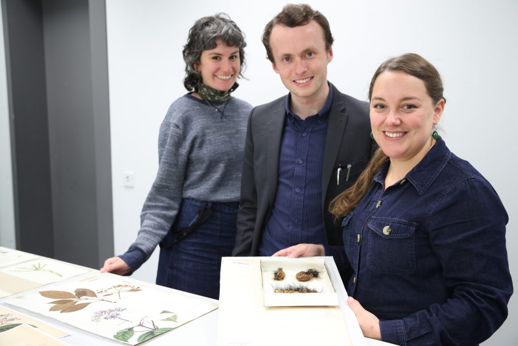 Two women and one man stand looking at the camera with one holding Darwin's cacti specimens, displayed on a piece of white card
