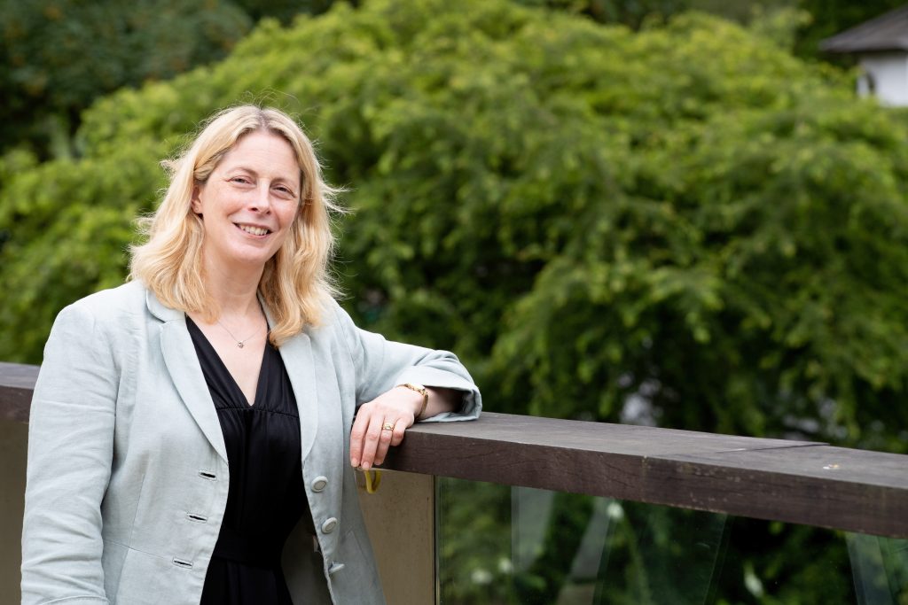 Professor Beverley Glover wearing a pale blue jacket and black top, leans against a balcony rail. A green tree is in the background.
