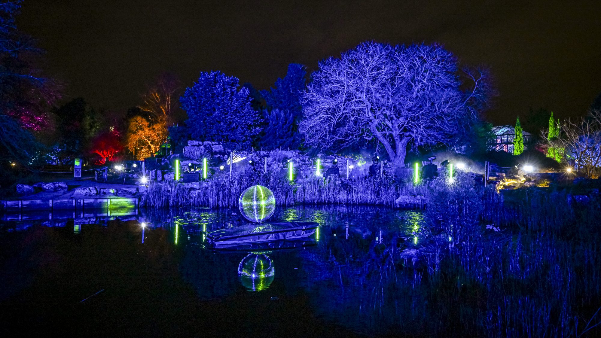 A night time scene of a lake bordered by trees lit up in different colours. A disco ball sits on a small boat in the lake and a glass building can be seen in the background.